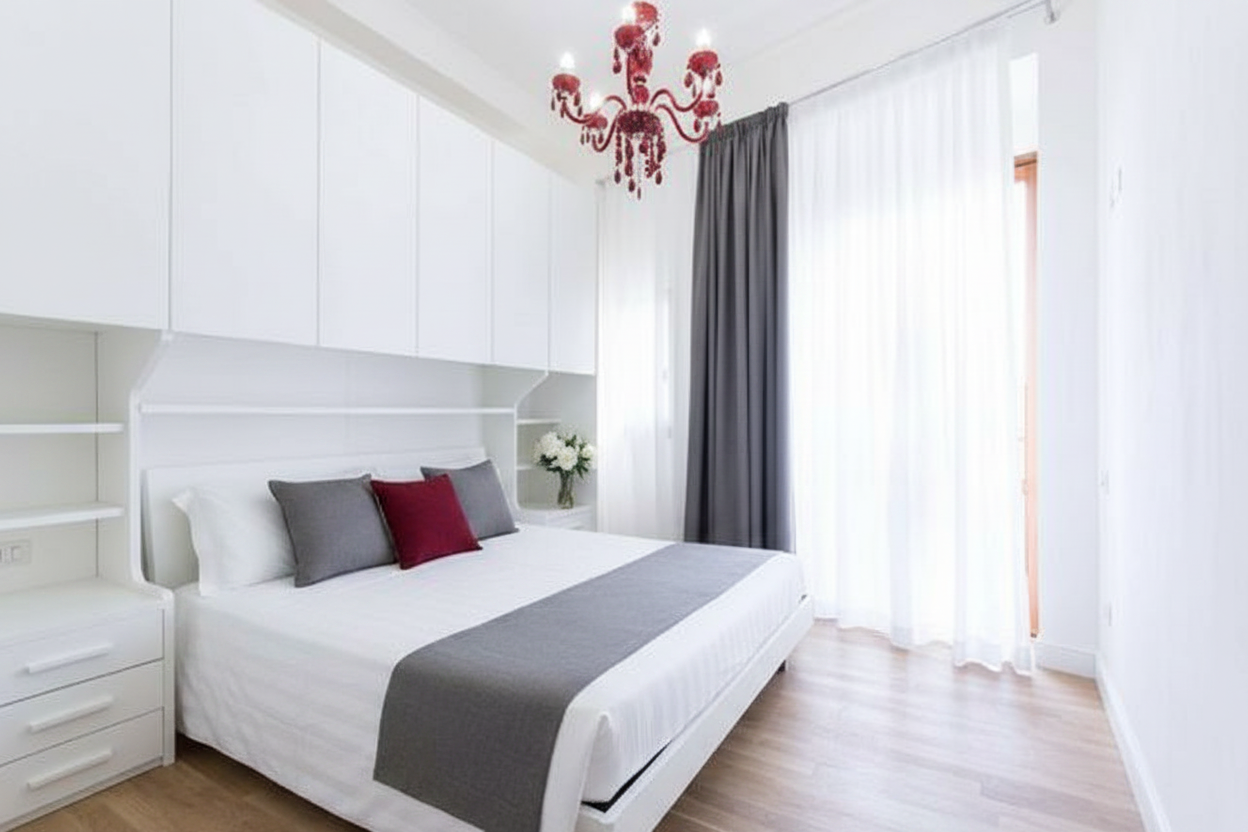 Exact same bedroom photo: white built-in wardrobes overhead, red Venetian glass chandelier, grey curtain on the right with natural light filtering through, light wood floor, white walls. The bed is a double bed with a white headboard integrated into the wardrobe system. Add: a crisp white hotel-style bedspread neatly tucked, with a folded grey runner/blanket draped across the foot of the bed. Three decorative pillows: two in grey matching the curtains, one accent pillow in deep red matching the chandelier. On the right nightstand, a small vase with white flowers (peonies or tulips). Keep the room architecture, furniture, chandelier and curtains exactly as they are — only add the bedding, pillows and flowers.